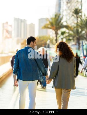 Young couple walks hand in hand on a lively city boardwalk under the sun, surrounded by modern buildings and palm trees in a warm, relaxed atmosphere. Stock Photo