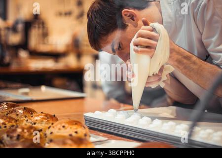Man in white chef's blouse piping meringue dessert using conic pastry bag with white creme. Stock Photo