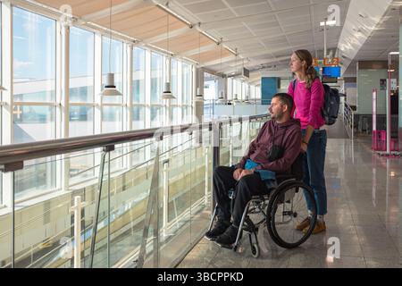 Man with disability using wheelchair with his female personal assistant is at the airport, looking out through large glass windows, preparing for Stock Photo