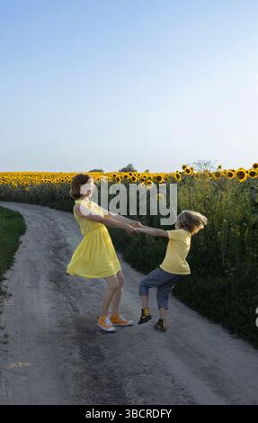 child and woman, mother and son holding hands and circling happily near sunflower blooming field. Happy childhood and joyful motherhood, having fun on Stock Photo