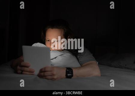 Man resting on bed, holding a tablet while browsing or reading in the dark with focused eyes. Stock Photo