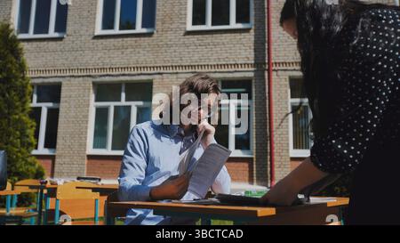 Two students are studying together at a desk in a schoolyard, preparing for exams on a sunny day Stock Photo