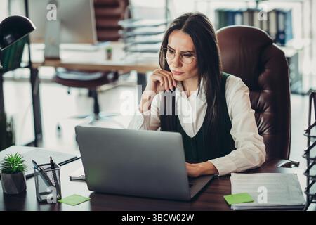 Experienced professional businesswoman focused on her work, analyzing documents and using a laptop in modern office interior Stock Photo