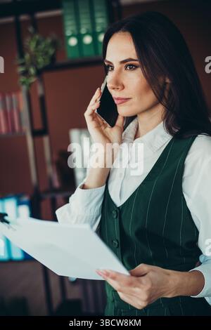Professional businesswoman discussing over the phone while holding documents in an elegant modern office setting Stock Photo