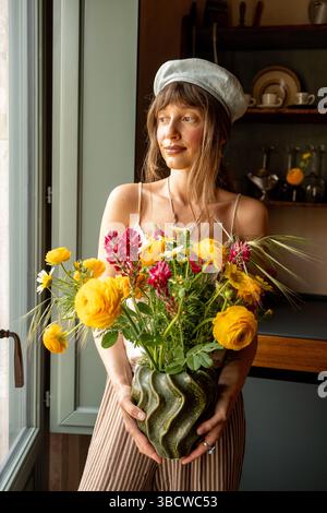 A young woman in a soft beret stands by the window holding a vibrant bouquet of yellow and magenta flowers, bathed in warm natural light. A peaceful, timeless scene of simplicity and charm Stock Photo