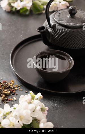 Black tea in cast iron teapot and cups decorated and white spring blossom flowers on black table. Japanese style and tea ceremony. Vertical Stock Photo