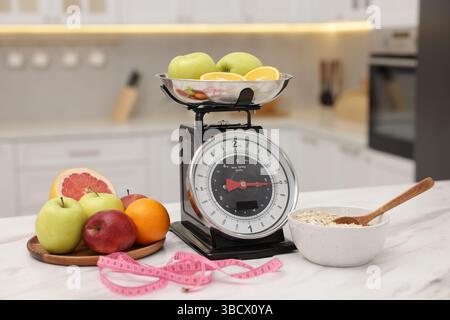 Kitchen scales, fruits, oatmeal and measuring tape on white marble table indoors Stock Photo