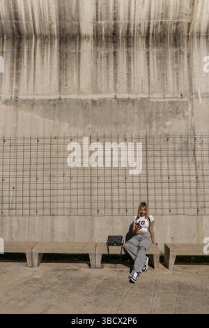 Fashionable young woman using a smartphone while taking a break on a concrete bench against a textured wall Stock Photo