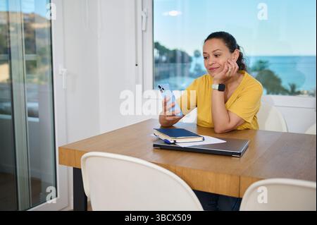 A woman sits at a desk in her home office, glancing thoughtfully at her smartphone. The room features a serene beach backdrop, emphasizing a balance b Stock Photo