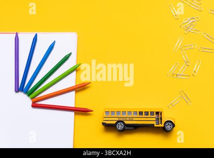 School supplies arranged on a yellow background, featuring colored pencils on a white notebook, scattered paper clips and a yellow toy school bus. Dep Stock Photo