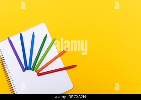 Colored crayons arranged in a fan shape on a white spiral notebook, placed on a bright yellow background. The image represents school, creativity, art Stock Photo
