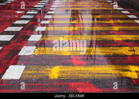 Colorful vivid pedestrian crosswalk with wet asphalt reflecting red, yellow, and white stripes during light snowfall and rain. Urban street, graphic Stock Photo