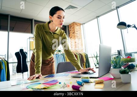 Young fashion designer working on laptop in a modern atelier surrounded by clothing and color palettes Stock Photo
