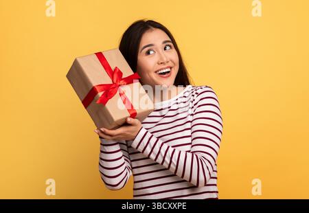Joyful woman holding a gift with a red ribbon against a bright yellow background Stock Photo
