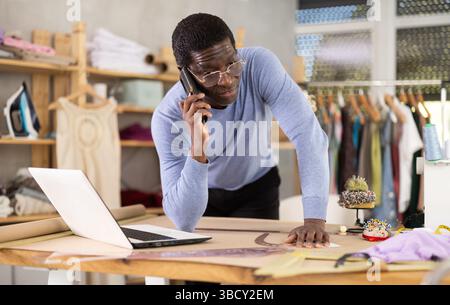 Tailor communicating on phone and reviewing specifications on laptop in workshop Stock Photo