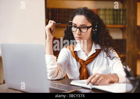 Focused woman sitting at desk, wearing headset, using computer Stock Photo