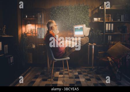 Middle-aged man working on an old computer in a cozy home office setting with casual style during the evening Stock Photo