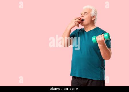 Sporty senior man exercising with dumbbell and eating apple on pink background Stock Photo