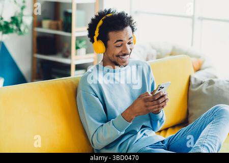 Smiling young man with headphones enjoying music on a yellow sofa in a cozy apartment during daylight Stock Photo
