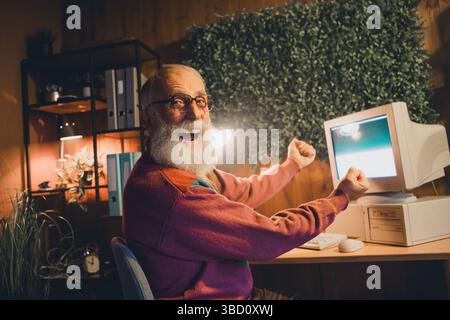 Elderly man joyfully working with a computer indoors in a cozy evening setting, showcasing freelance programming dedication and skill Stock Photo