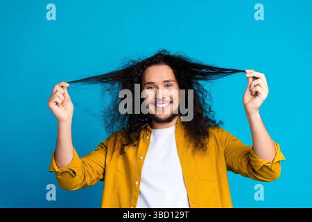 Cheerful man with curly hair in yellow shirt smiling against vibrant blue studio background Stock Photo