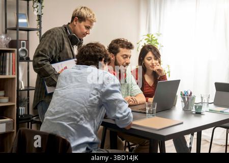 Smiling Gen-Z coworkers cooperate on a design project, analysing data on laptop in casual home office; concept of teamwork, innovation and hybrid work Stock Photo