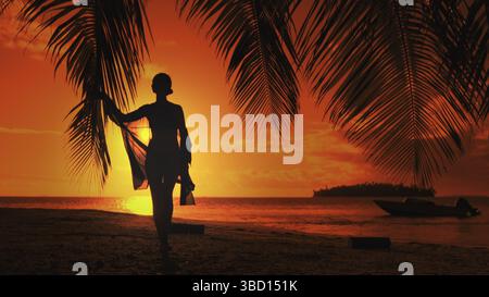 Woman walks gracefully on a tropical beach at sunset, holding a palm frond, gazing at the ocean. The sky is orange, red, and yellow, creating a romantic atmosphere Stock Photo