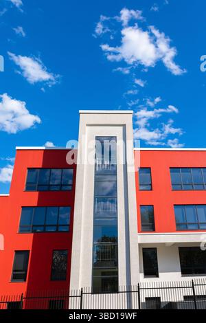 Vertical view of a modern red and white building with large glass windows and a clear blue sky with scattered clouds. Stock Photo