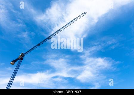 Tall blue construction crane extending diagonally across a bright sky with scattered white clouds. Stock Photo