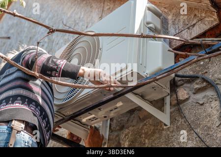 A man installs an outdoor air conditioner unit on iron mounting brackets on the wall of a stone rural house. DIY installation. Stock Photo