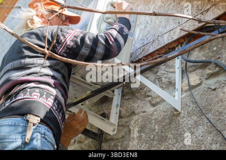 A man installs an outdoor air conditioner unit on iron mounting brackets on the wall of a stone rural house. DIY installation. Stock Photo