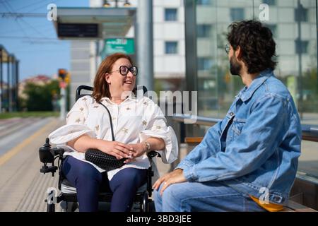 Disabled woman in wheelchair smiling and talking with man at tram station - wheelchair and disability concept Stock Photo