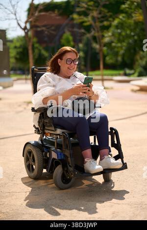 Smiling woman using smartphone while sitting in electric wheelchair in park - wheelchair and disability concept Stock Photo