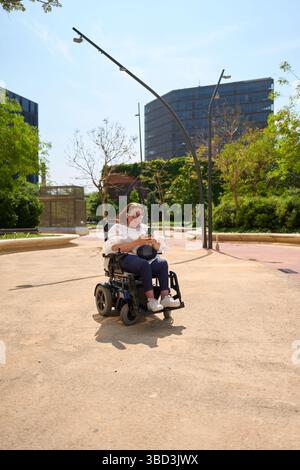 Smiling woman using smartphone while sitting on wheelchair in park - wheelchair and disability concept Stock Photo