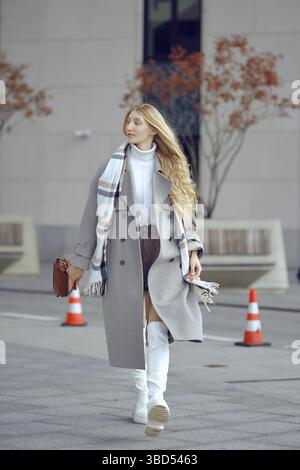 A fashionable woman walks along a city street, wearing a stylish gray coat and knee-high white boots. She carries a small handbag and is surrounded by Stock Photo