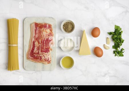 Fresh ingredients on a marble countertop for cooking spaghetti carbonara. Includes bacon, eggs, cheese, garlic, herbs, black pepper, and olive oil, re Stock Photo