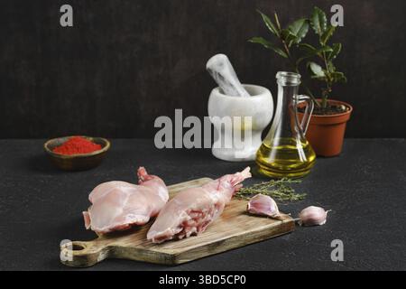Fresh rabbit legs on a wooden cutting board alongside spices, garlic, and herbs. An olive oil bottle and a mortar with pestle add to the cooking setup Stock Photo