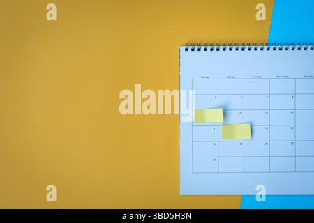 Calendar page with yellow adhesive notes. On top of yellow and blue surface, high angle view. Copy space. Stock Photo