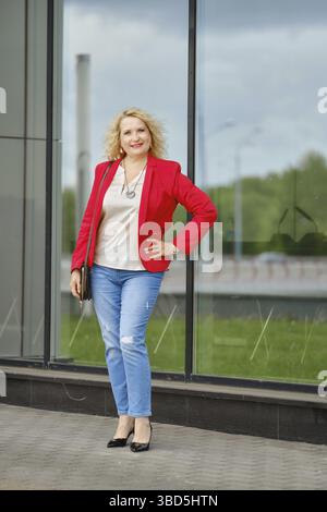 Confident woman in red jacket, blouse and denim jeans standing near the reinforced glass wall of the building Stock Photo
