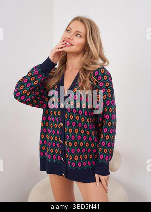 Blonde woman with curly hair in colorful patterned sweater, smiling with hand by face. Sweater features vibrant red, blue, and yellow designs. Casual Stock Photo