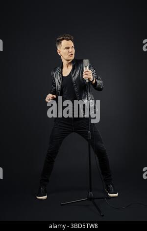 Man in black leather jacket, jeans and shirt with microphone in hand over dark background Stock Photo
