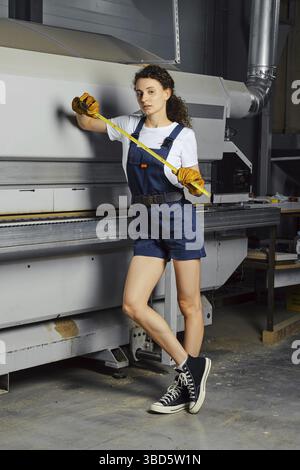 Apprentice woman leaned against a woodworking machine with a steel tape measure in hand Stock Photo