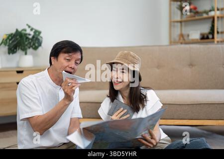Elderly Asian couple joyfully exploring a travel map and discussing plans while holding a passport, highlighting travel excitement. Stock Photo