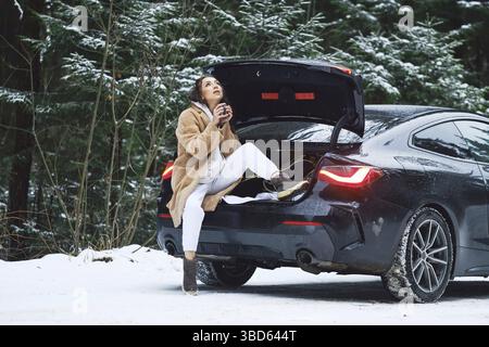 Tired driver woman stopped her car in winter forest road to give herself rest and to drink a cup of coffee Stock Photo