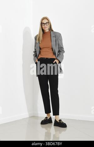 Stylish woman wearing checkered blazer and brown turtle neck sweater and black trousers standing against white office wall Stock Photo