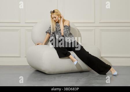 A beautiful blond-haired woman in fashionable boho style clothes sits with her legs crossed in a soft bean bag chair. Her loose trousers and stiletto Stock Photo