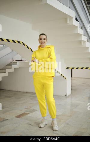 A woman stands on a staircase wearing a bright yellow hoodie and matching pants. She smiles confidently against a backdrop of clean lines and contempo Stock Photo