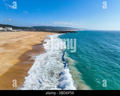 Clear blue wave rolling onto a wide sandy beach with a few people walking nearby Stock Photo