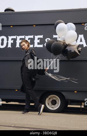 Pretty young woman in black outfit with balloons in hand walking on the street Stock Photo
