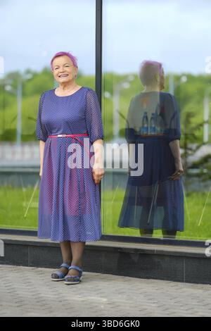 Old woman with cheerful smile in blue dress standing near the modern building with glass walls Stock Photo